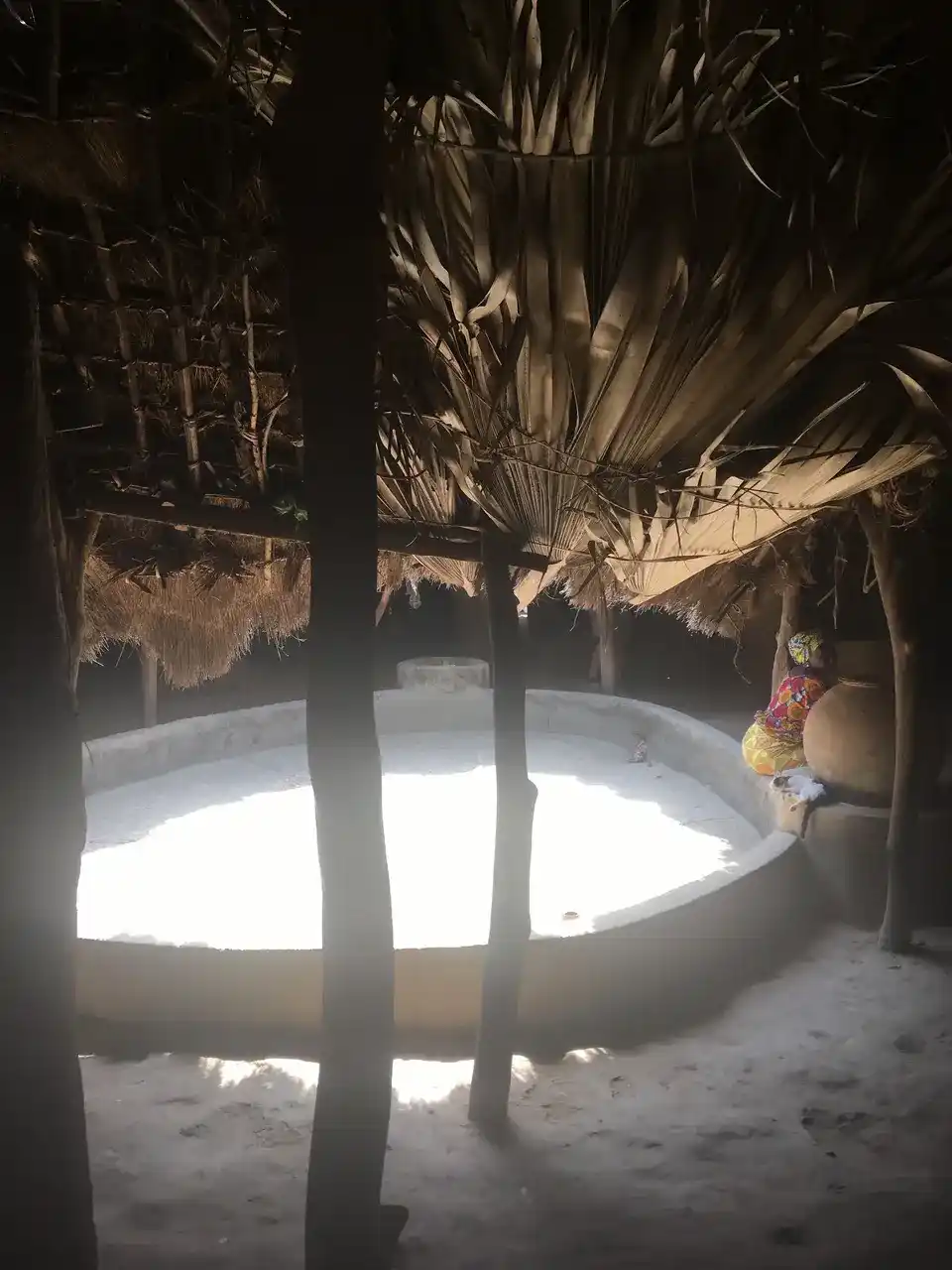 Le bassin central de la case à impluvium d'Enampore, éclairé par l'ouverture du toit, avec une jarre en terre et une femme assise au fond