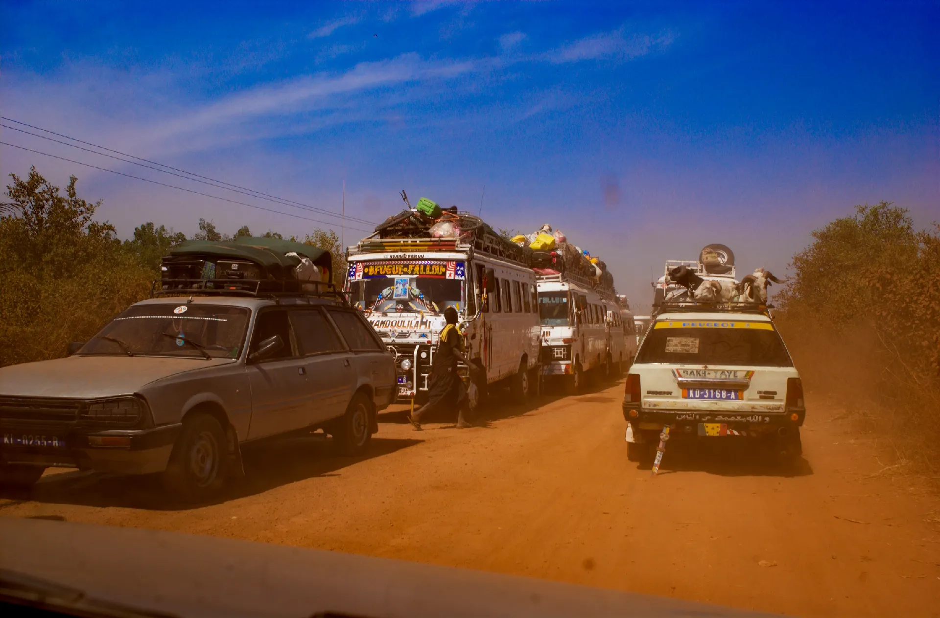File de taxis-brousse et de cars rapides surchargés sur une piste de latérite poussiéreuse